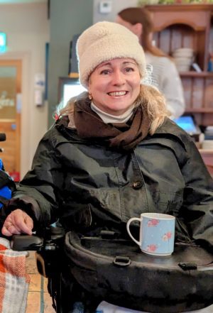 lady in a white bobble hat smiling with a cup on a trabasack wheelchair tray, she is seated in a power chair