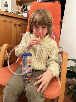 A young boy with blonde hair sits in an orange chair, using a HydroBubble device. He is wearing a cream-colored sweater and holding a clear container painted with colorful bubbles connected to a tube that he is breathing into. The setting appears to be a cozy indoor space with wooden furniture and some plants.