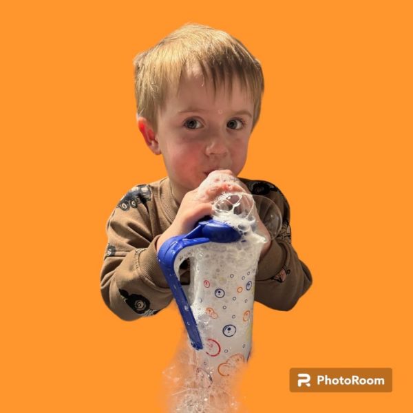 A young boy with light brown hair uses a HydroBubble device against an orange background. He is wearing a brown shirt with black patterns and holding a white container with colorful designs connected to a blue tube that he is breathing into