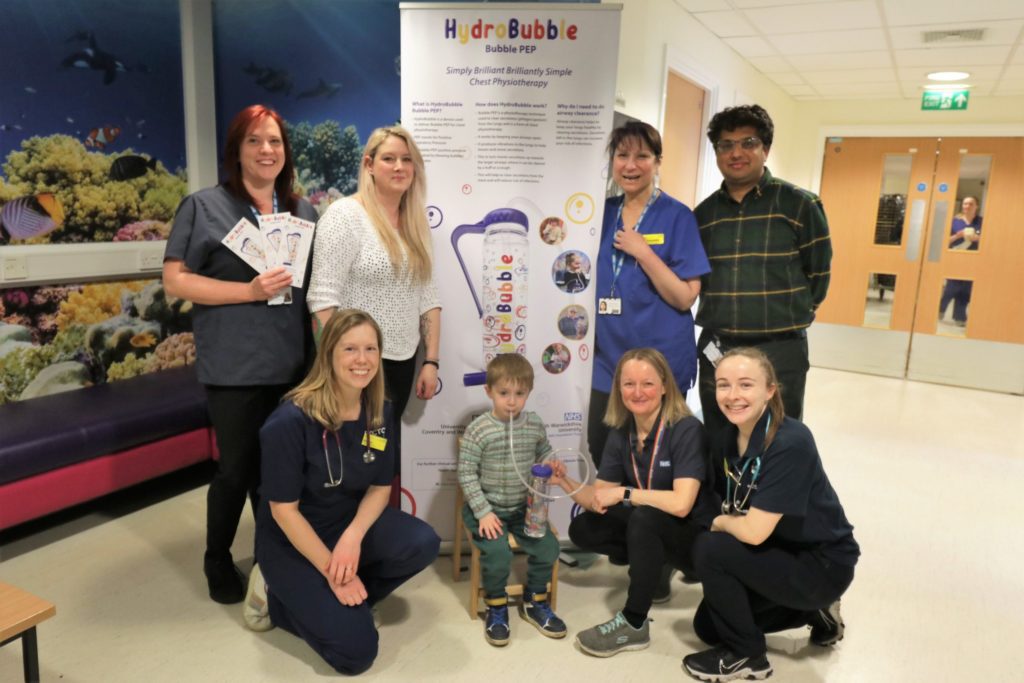 A group of healthcare workers, including five women and one man, pose with a young boy in front of a poster for "HydroBubble Bubble PEP" in a hospital hallway. The boy holds a Bubble PEP device, and the workers are smiling, wearing various uniforms. The background features an aquarium-themed wall.