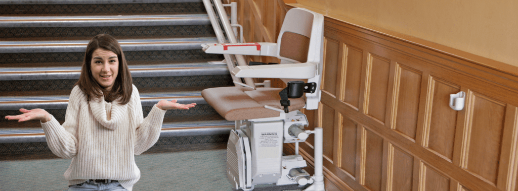 A woman with a puzzled expression stands at the base of a staircase, gesturing with both hands. To her right is a stairlift attached to the wall, positioned next to the stairs. The setting appears to be indoors with wooden paneling on the wall and carpeted steps.