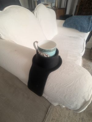 A ceramic mug is placed securely in a black CouchCoaster, which is draped over the armrest of a white sofa. The CouchCoaster prevents spills by holding the mug steady on the curved surface. The room has soft lighting, and a blue throw blanket is seen on the sofa in the background.