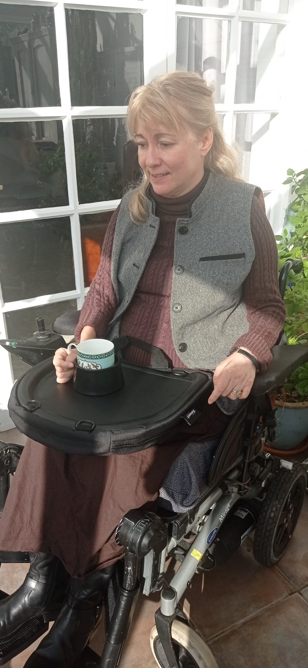 A woman in a wheelchair holds a mug resting on a black lap tray, which is attached to her wheelchair. The tray securely holds the mug using a cup holder attachment. She is dressed in a maroon sweater and a grey vest, seated indoors by a glass door, with sunlight streaming in.