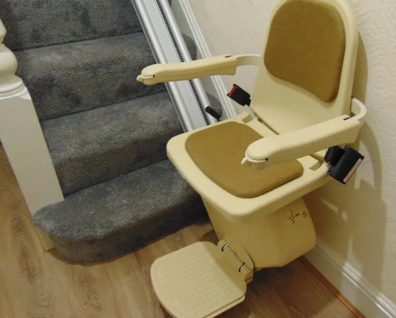 A stairlift installed beside a carpeted staircase. It has a padded beige seat with armrests, a footrest, and safety belts. The chair is positioned at the base of the stairs, ready to transport a person up and down safely, providing a practical solution for older adults or those with mobility issues.