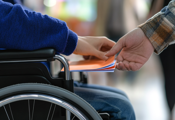 A person seated in a wheelchair receives a stack of orange and white papers from someone standing nearby. The setting is softly blurred, suggesting an indoor environment.