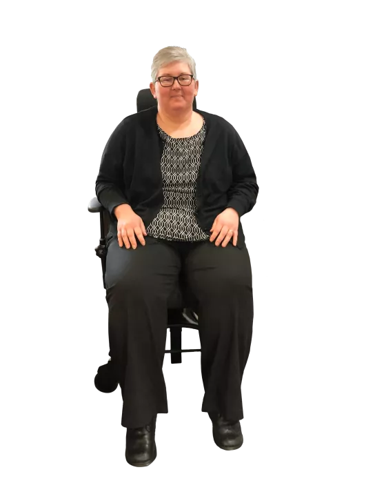 A woman with short gray hair and glasses is seated on a office chair with wheels, facing forward. She is wearing a patterned top, a black cardigan, black pants, and black shoes. The background is transparent.