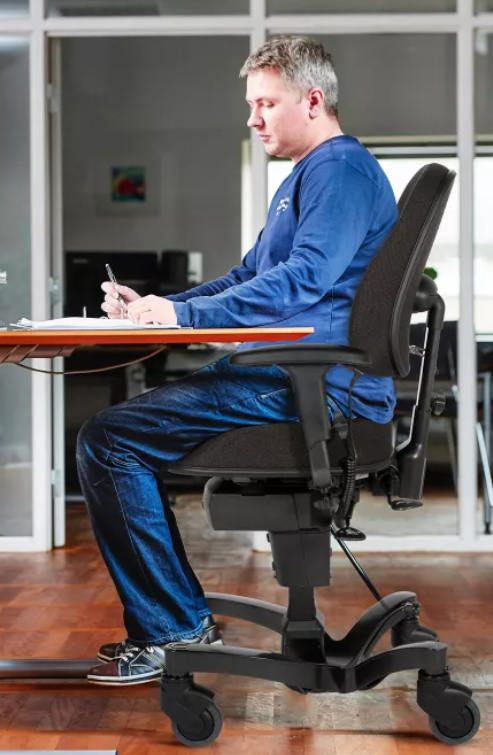 A man in a blue long-sleeve shirt and jeans sits upright on a wheeled office chair with brakes at a desk, writing on paper. The chair has an ergonomic design with adjustable support and wheels. The setting is an office with glass walls and wooden flooring.