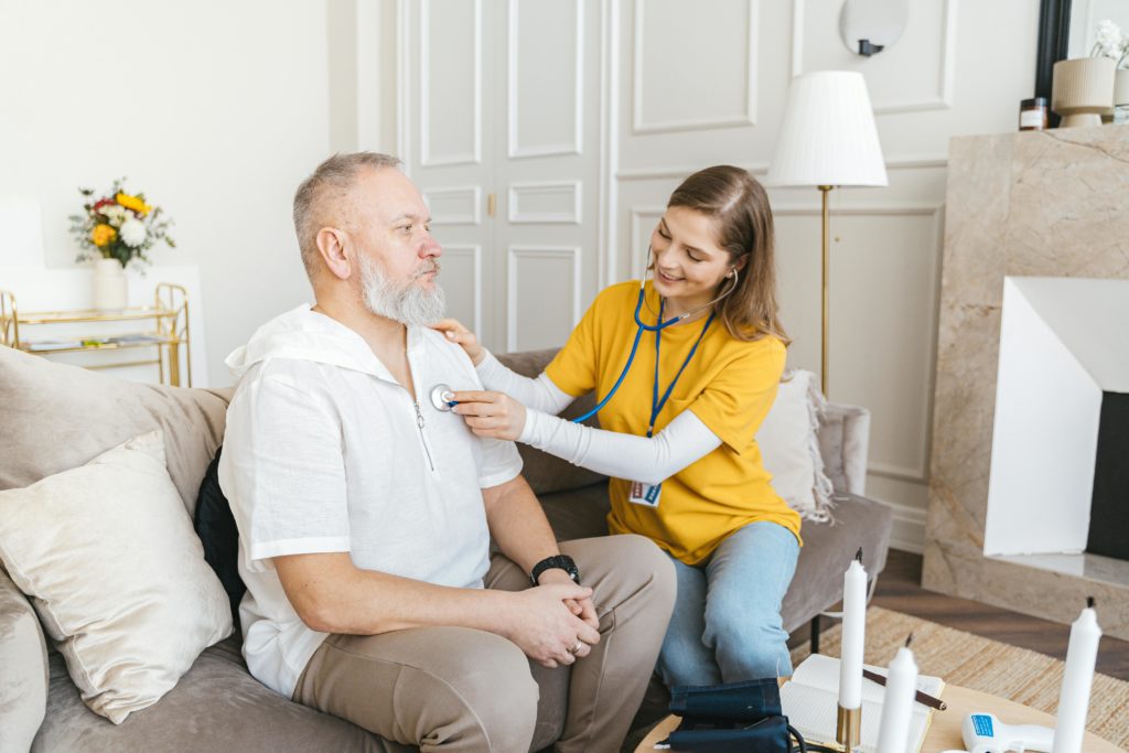 A young female caregiver in a yellow shirt and blue jeans is using a stethoscope to check the heartbeat of an older man with a white beard, who is sitting on a beige couch. The man is wearing a white shirt and brown pants, looking straight ahead. The setting is a well-lit, modern living room with a fireplace, a coffee table with medical tools, and a vase with flowers in the background.