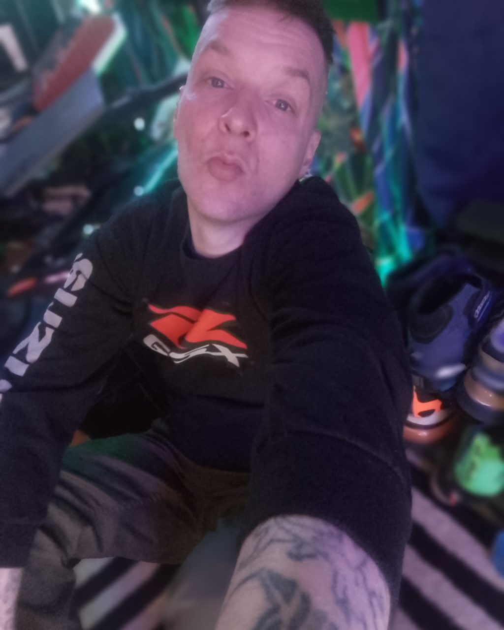 Joseph with short hair and tattoos on his arm is taking a close-up selfie, puckering their lips. They are wearing a black long-sleeve shirt with red and white graphics. The background is colorful and slightly blurred, featuring various objects, including shoes and neon lighting.