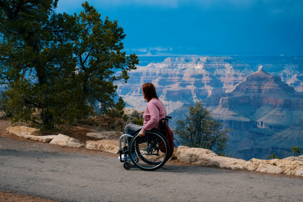 A woman in a wheelchair, wearing a pink sweater and gray pants, gazes at the Grand Canyon from a paved overlook. The canyon’s layered rock formations stretch into the distance under a cloudy blue sky, with trees and boulders framing the scene.