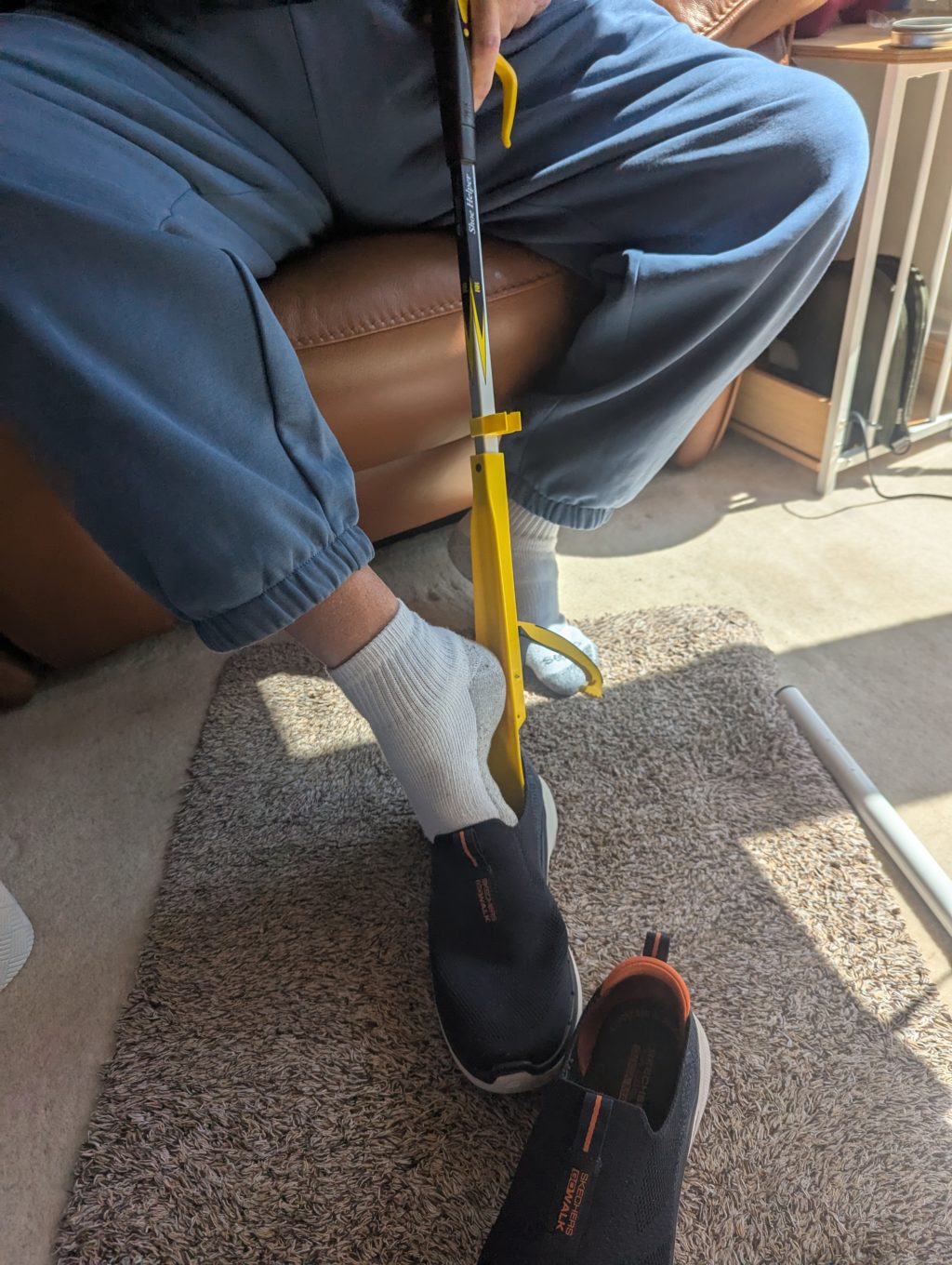 A person uses a yellow reacher tool to help put on a black slip-on shoe while seated.