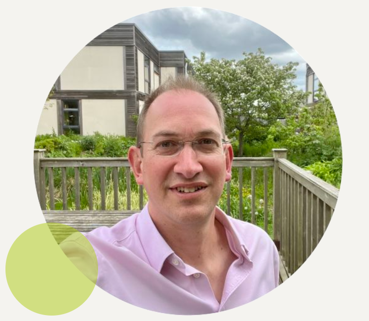 Guy Harris wearing glasses and a light pink shirt standing on a wooden deck with greenery and a modern building in the background.