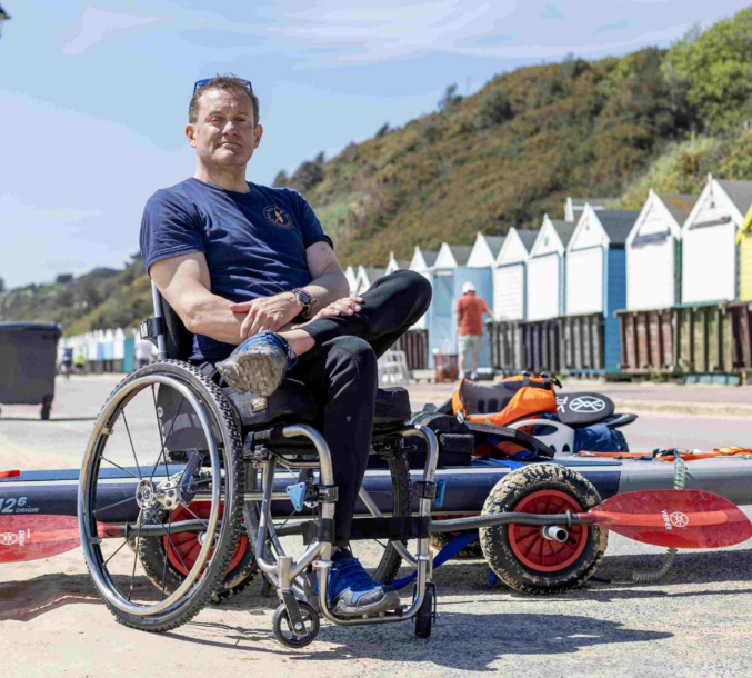 Will in a wheelchair sitting beside a paddleboard and beach cart, with colorful beach huts and cliffs in the background.