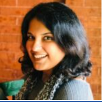 Hema Thakur, a smiling woman with shoulder-length dark hair, wearing a grey top and scarf, sitting against a brick wall background.