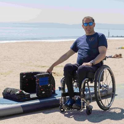 Will Behenna in a wheelchair on a beach beside an adapted paddleboard with a foam seat, smiling and pointing to the equipment.