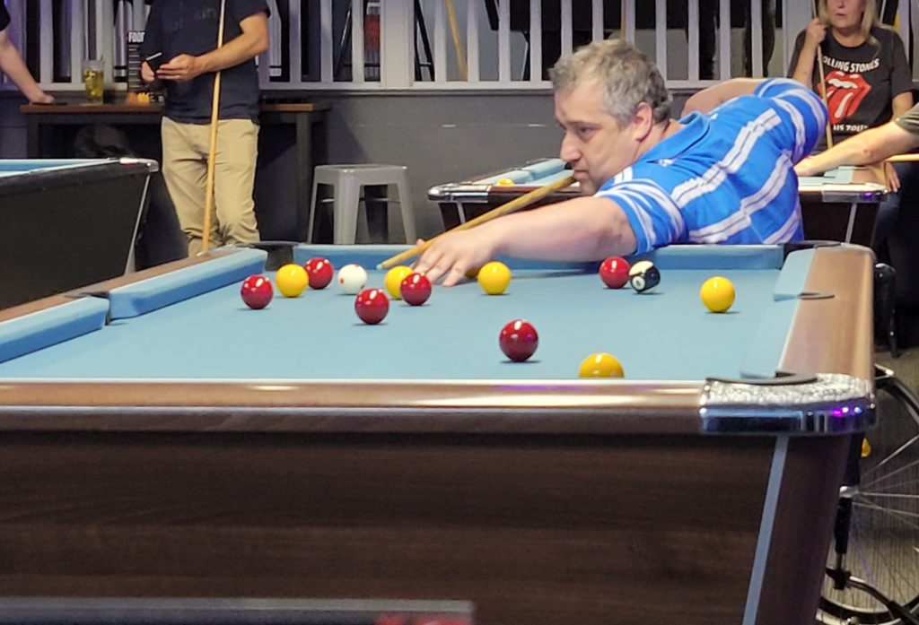 Gareth in a manual wheelchair wearing a blue striped shirt, leans forward with focus as he lines up a pool shot. The table is crowded with red, yellow, and black balls. Other players stand nearby, watching or waiting their turn.