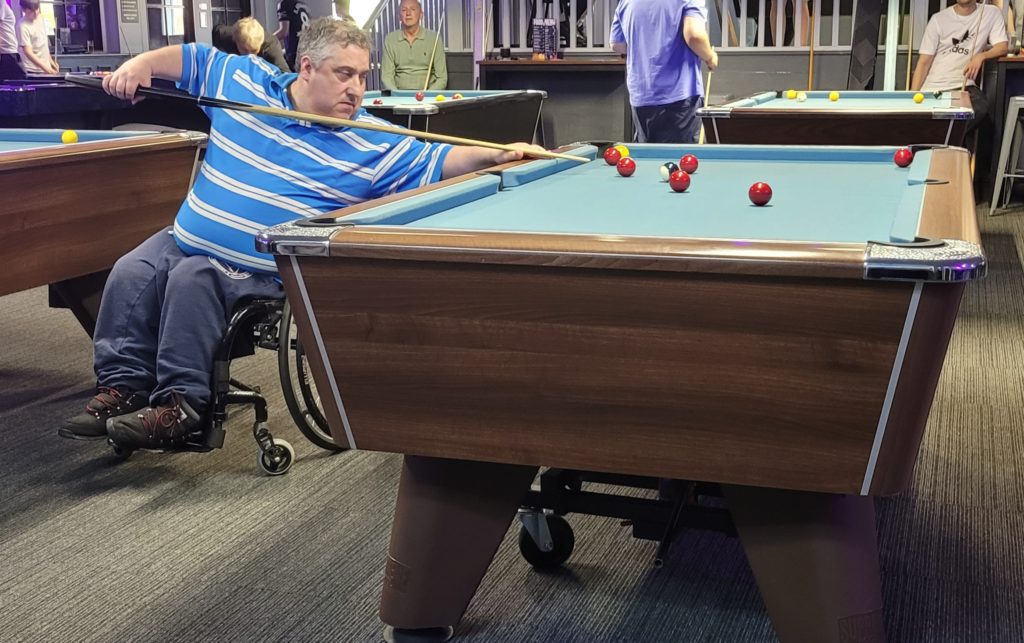 Gareth Clarke A man in a wheelchair, wearing a blue striped shirt, is focused and mid-shot during a game of pool. He stretches across the table to line up his cue, aiming at a cluster of red and yellow balls. Several other players and pool tables are visible in the background.