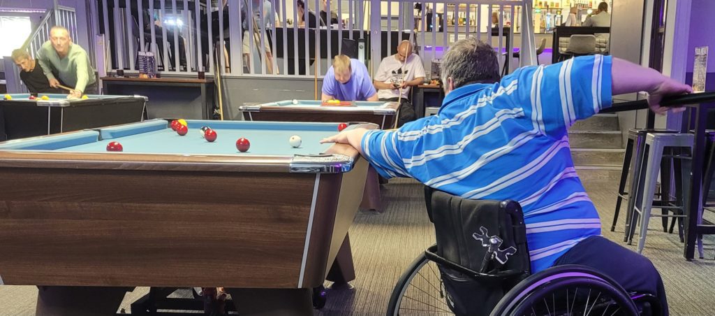 Gareth, a competitive wheelchair cue sports player, wearing a blue striped shirt, prepares to take a shot at a pool table. Several red and yellow balls are scattered across the table. Other players are visible in the background at nearby tables, and a bar area is seen further back.