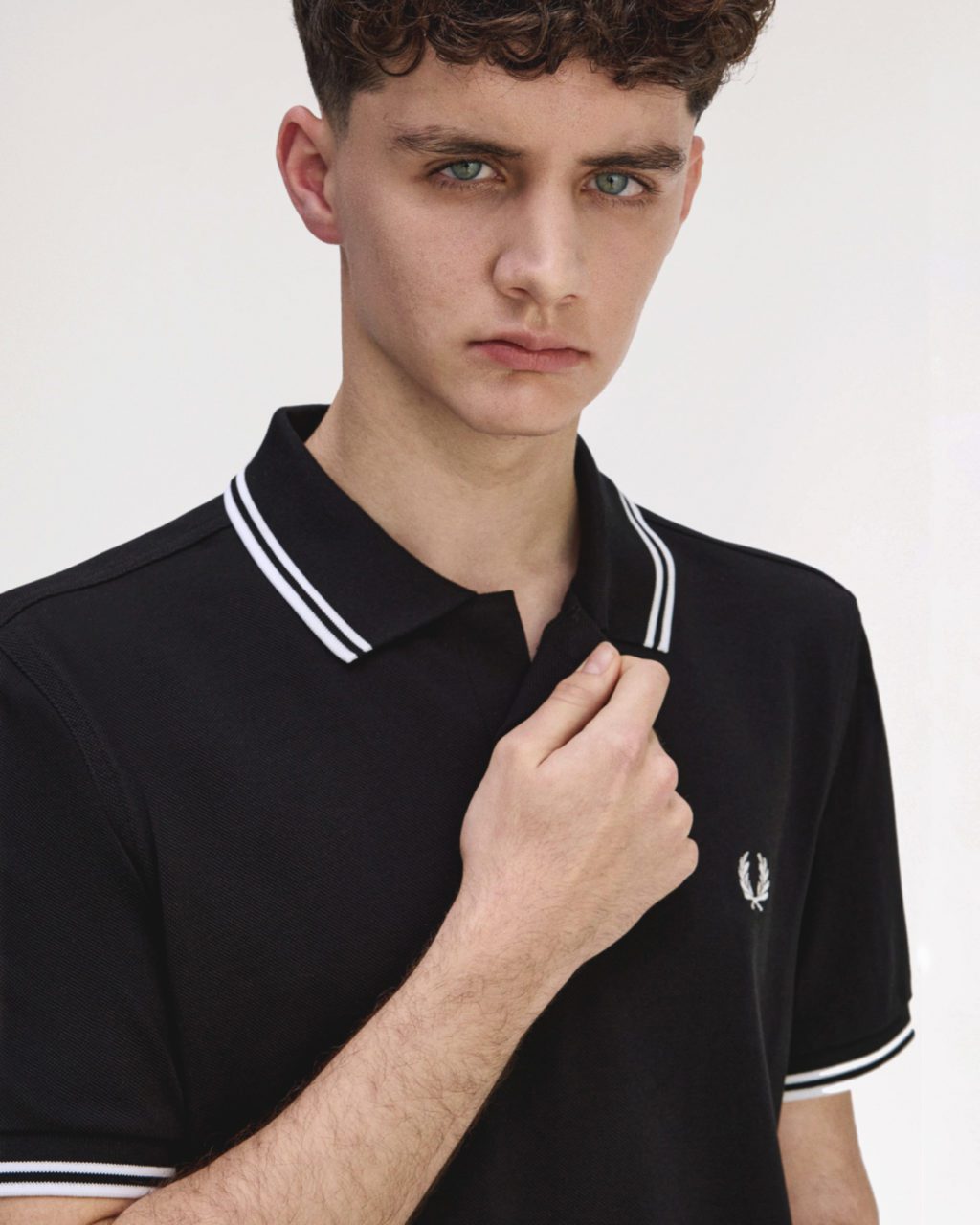 Young man wearing a black Fred Perry adaptive polo shirt, holding the collar to show its magnetic fastening.