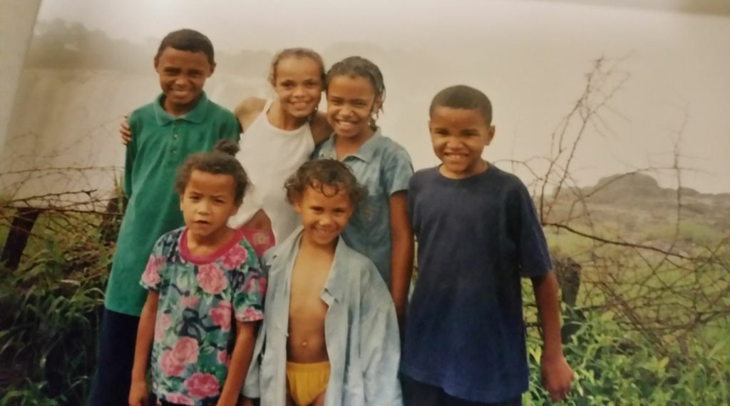 Six children smile and pose together outdoors near a misty waterfall and barbed wire fence. Their clothes are casual and wet, suggesting they’ve been playing in water. The background includes lush greenery and fog.