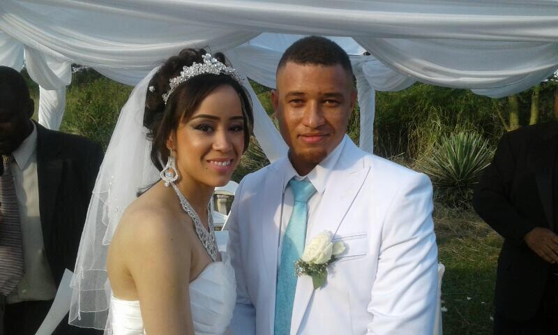 Bride and groom posing outdoors under white drapery. The bride wears a strapless white gown, tiara, and veil; the groom wears a white suit with a light blue tie and boutonnière.