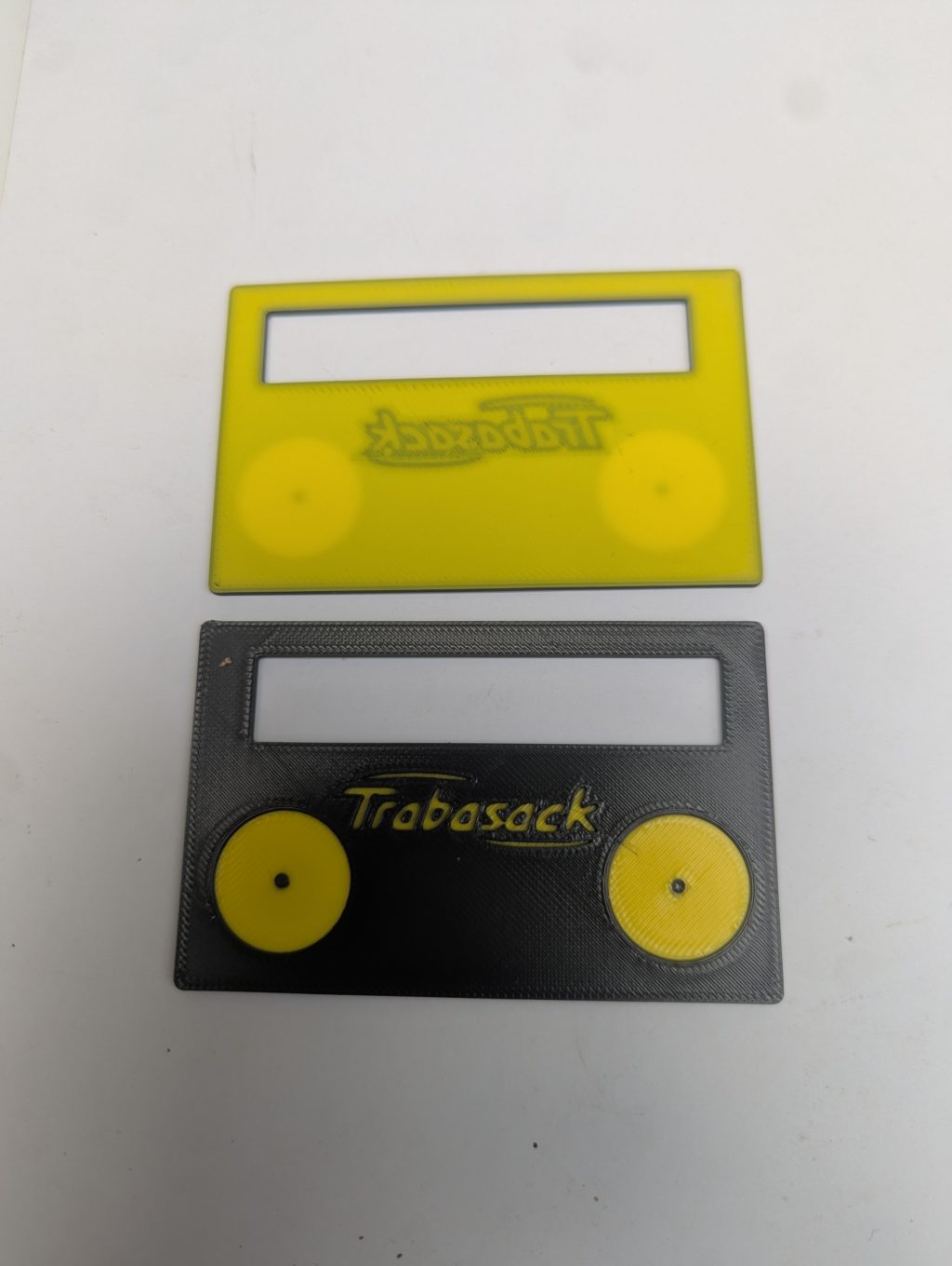 Credit-card–sized yellow and black plastic signature guide with a rectangular viewing window at the top and two circular thumb rests, labeled “Trabasack.”