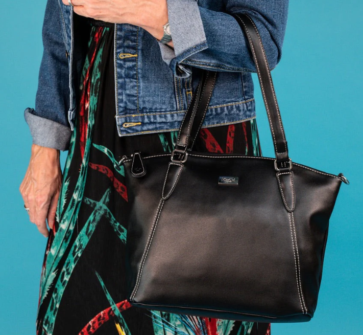 A person wearing a denim jacket and patterned dress holds a black Samantha Renke handbag with white stitching and two shoulder straps, against a blue background.