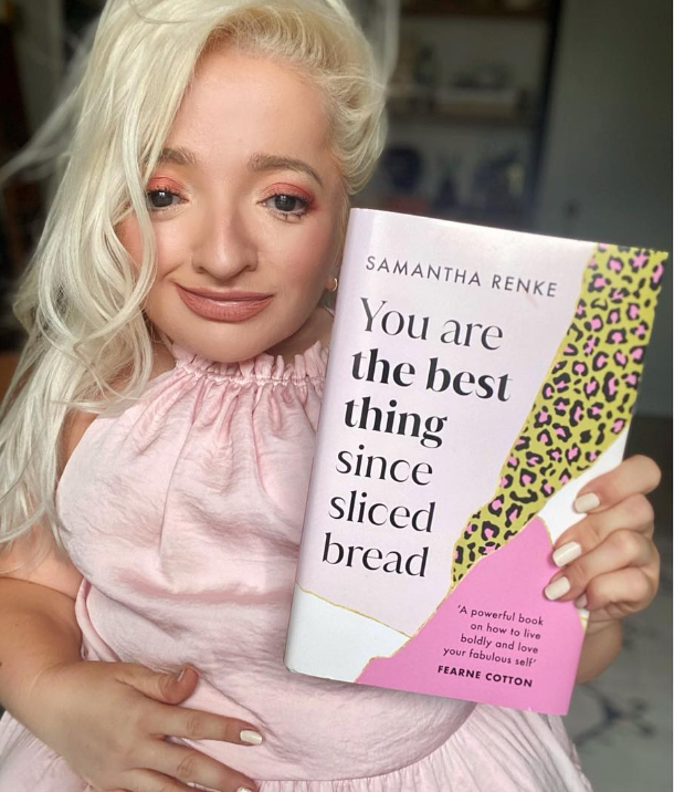 Samantha Renke holds her book titled You Are the Best Thing Since Sliced Bread. She is wearing a light pink, sleeveless ruffled top and smiling slightly. The book cover is pink, white, and yellow with leopard print accents and includes a quote from Fearne Cotton
