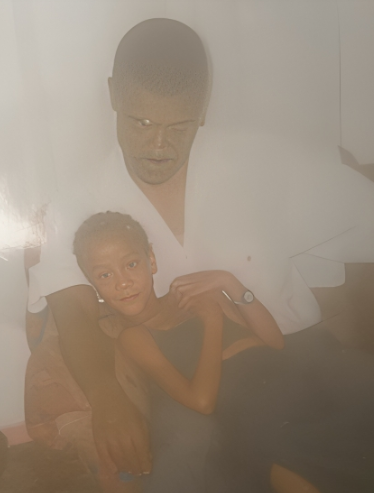 A man sits on a couch holding a young girl who is lying across his lap. The lighting is dim and the image appears faded or overexposed.
