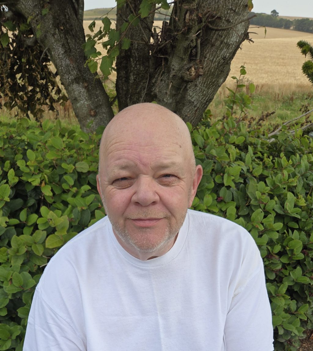 Eddie Thomson in a white shirt sitting outdoors in front of green bushes and a tree, with golden fields in the background.