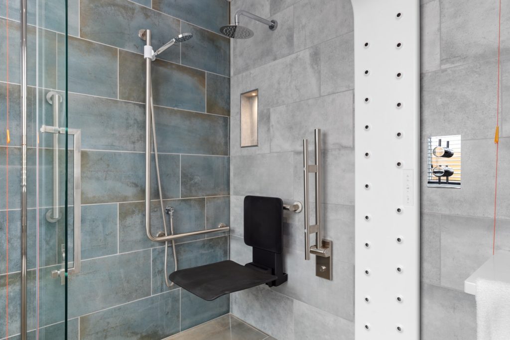 Modern accessible shower with a fold-down seat, grab bars, handheld and overhead showerheads, and a vertical panel of body air dryers.