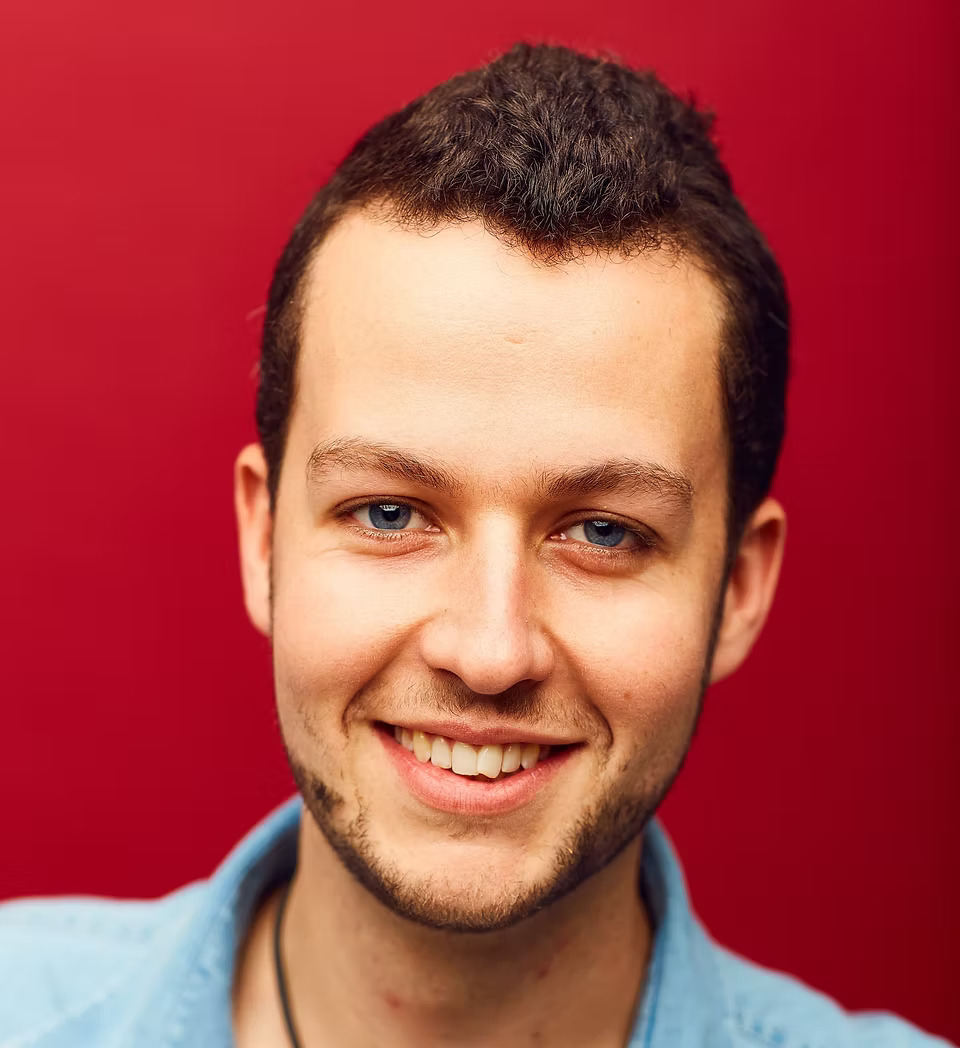 Dave Chawdner, young 30ish man with slight beard growth on a red background, he is smiling and wears a blue shirt