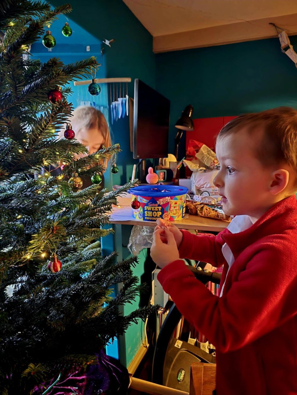 A young child in a red jacket decorates a Christmas tree with shiny baubles. Another child is partially visible behind the tree. The room is cozy and cluttered, with a TV, lamp, and snacks on a table, including a colorful “Sweet Shop” tub.