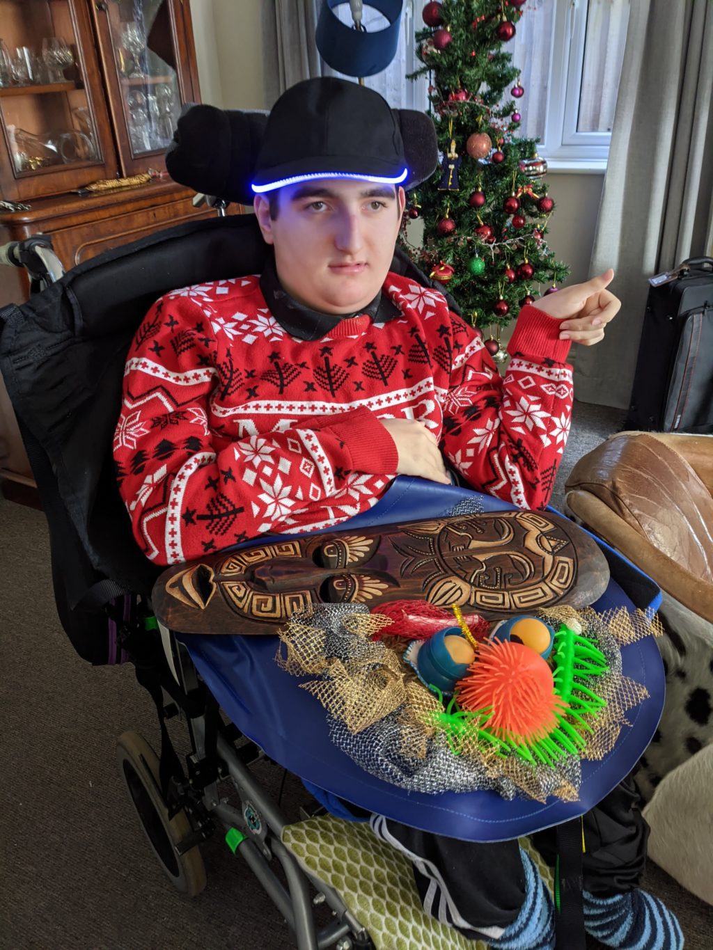A young man in a wheelchair wears a red festive sweater and a black cap with a glowing blue rim. He sits in front of a decorated Christmas tree, with sensory toys and a carved wooden mask on a trabasack tray across his lap.