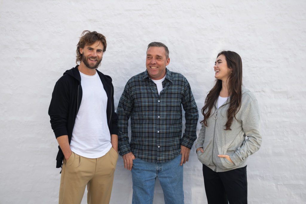 Three adults in casual shirts and zip-up hoodie stand against a white wall, smiling and chatting.