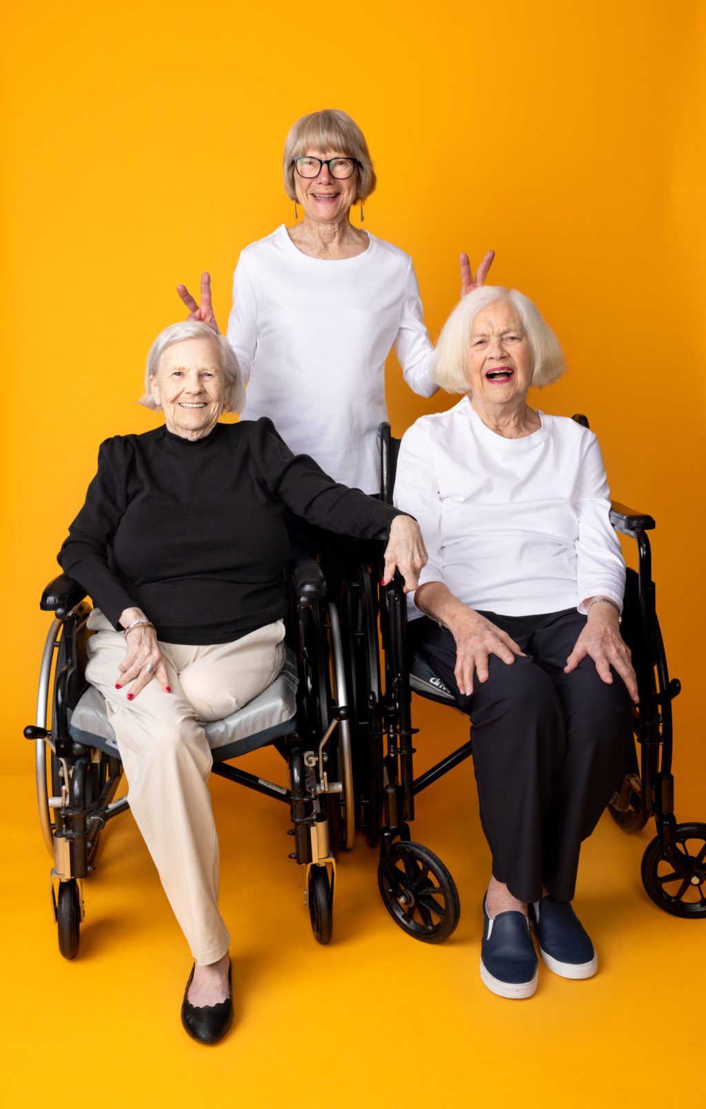 Three older women wearing simple, comfortable outfits smile against a bright orange background, two seated in wheelchairs and one standing behind them.
