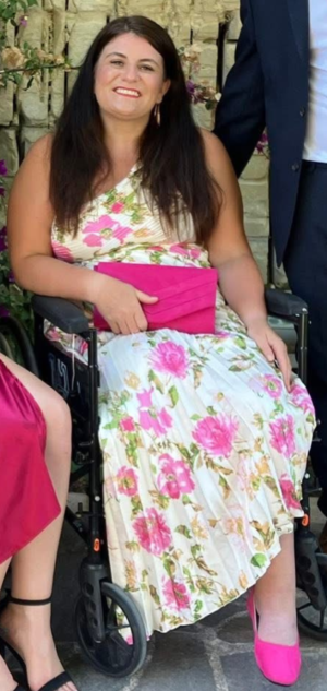 Sarah Beamis smiling in a wheelchair, wearing a white floral dress and holding a pink clutch outdoors.
