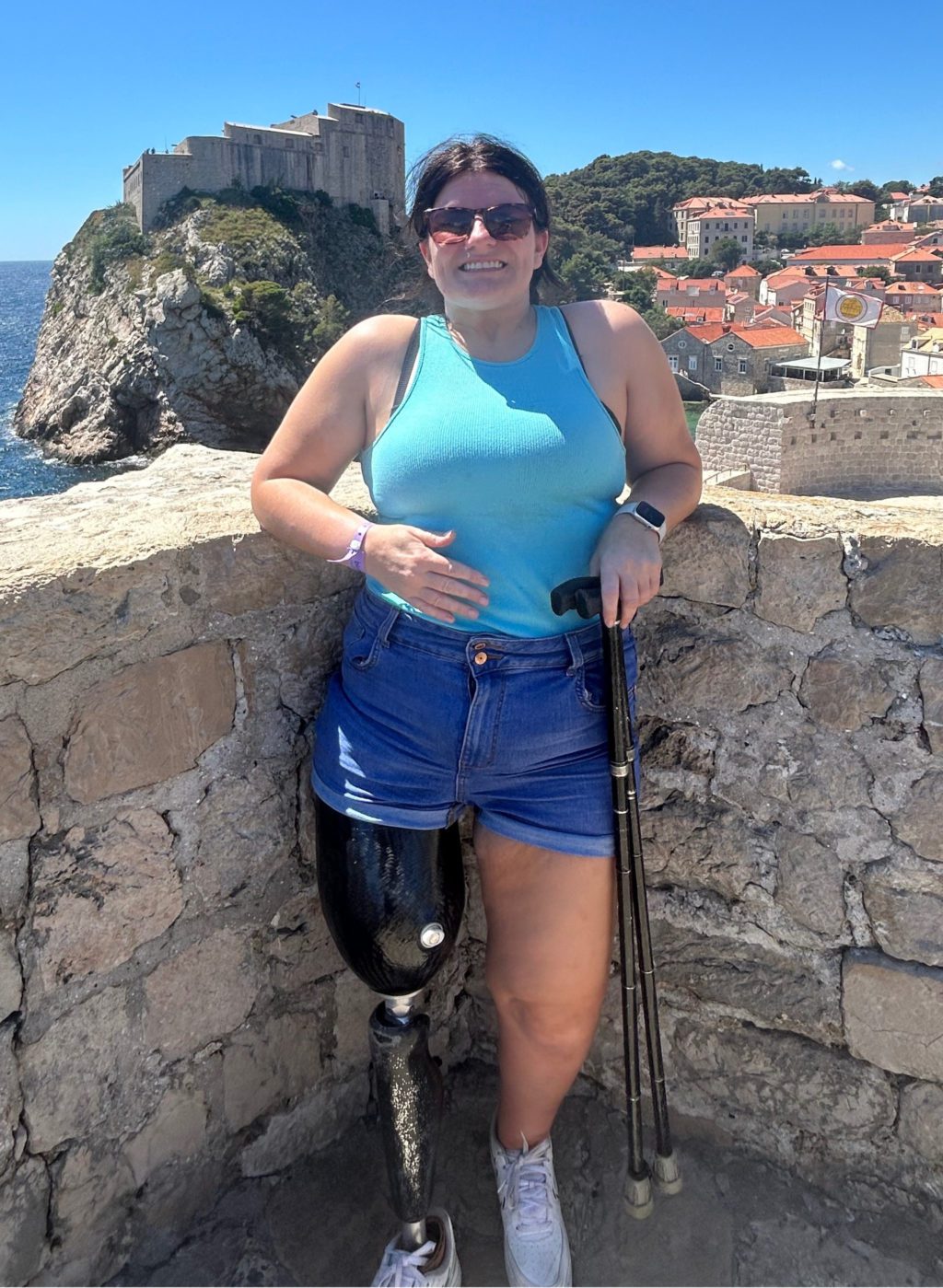 Sarah Beamis smiling outdoors by a coastal fort, wearing a leg prosthesis and holding crutches.
