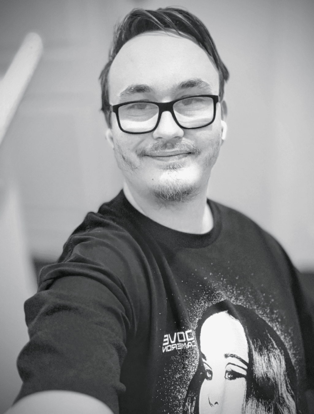 Black-and-white selfie of a smiling person with glasses and earbuds, wearing a Dove Cameron sweatshirt.