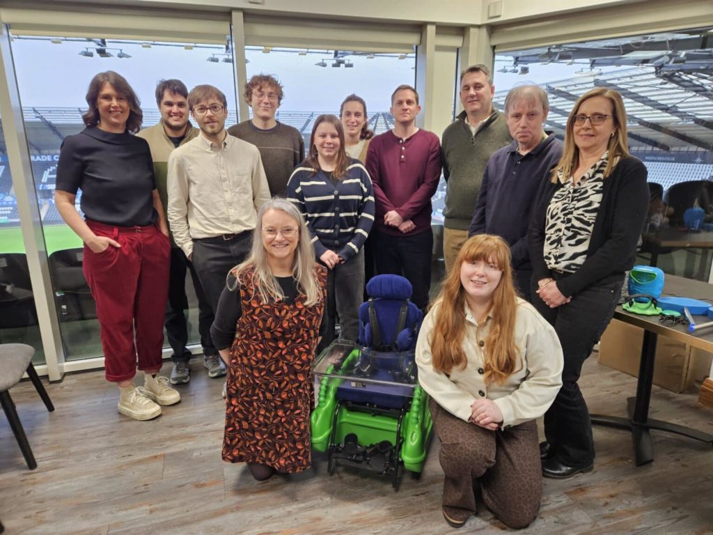 Team from Cerebra and MERU standing together with a Bugzi powered wheelchair in a meeting room overlooking a stadium.