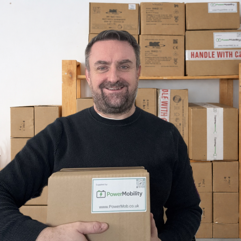 Tim smiling while holding a cardboard box labeled “PowerMobility, www.PowerMob.co.uk,” with stacked shipping boxes behind him.