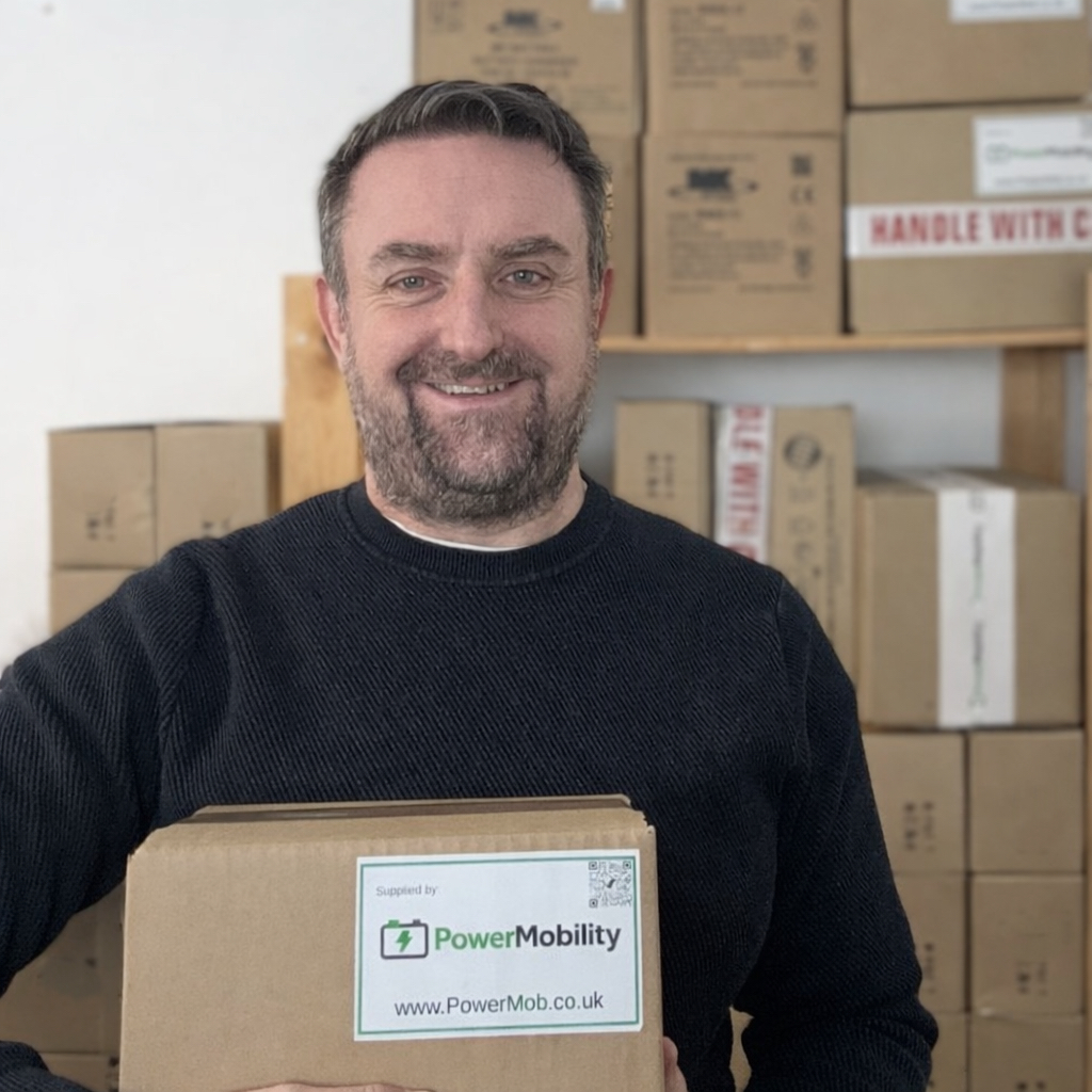 Man smiling while holding a box labeled “PowerMobility, www.PowerMob.co.uk,” standing in front of stacked shipping boxes.