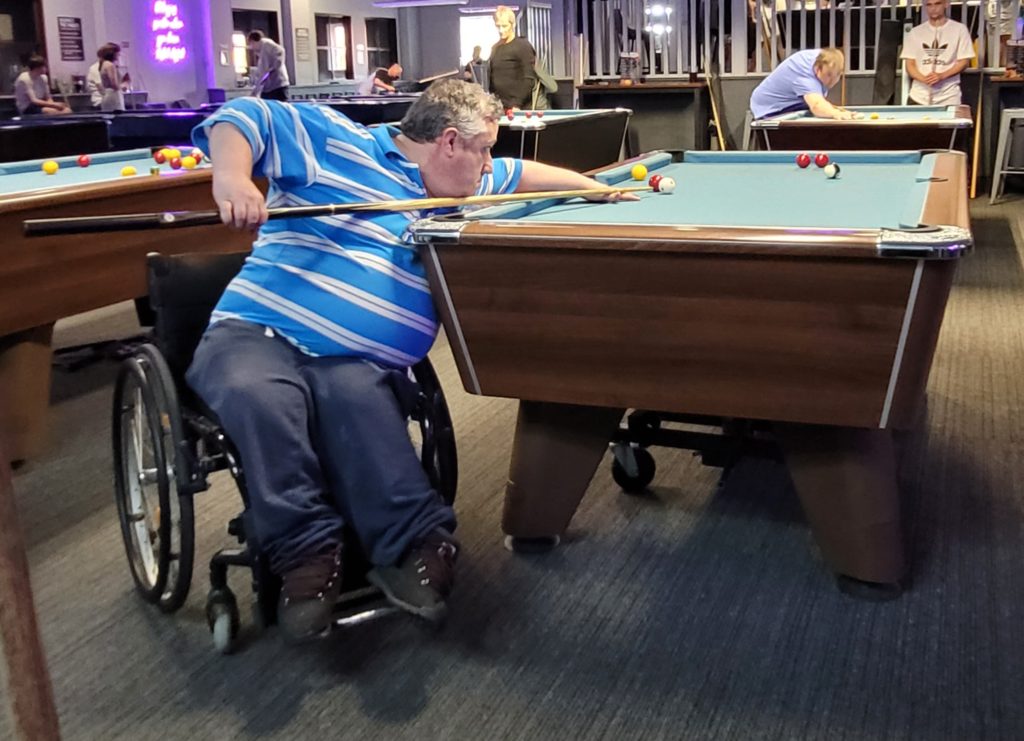 Gareth in a manual wheelchair leans across a pool table to line up a shot in a busy billiards hall.