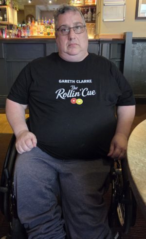Gareth wearing glasses sits in a wheelchair at a pub table, wearing a black T-shirt reading "GARETH CLARKE The Rollin' Cue" with a cue ball logo.