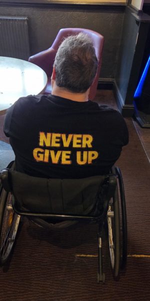 Rear view of a Gareth in a manual wheelchair wearing a black T-shirt with the words "NEVER GIVE UP" in large yellow letters.